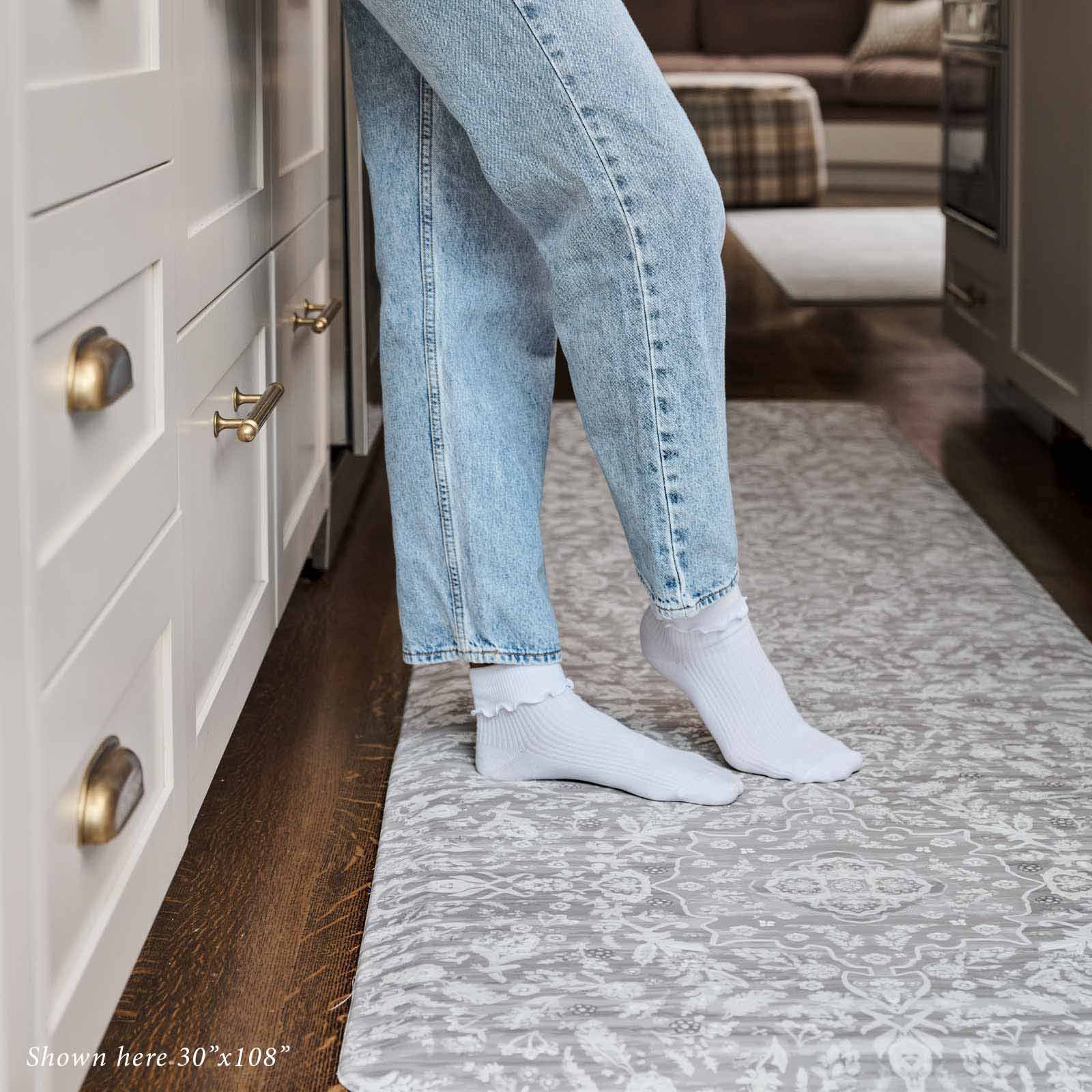 Emile earl grey gray and cream floral standing mat shown in kitchen in size 30x108 with womans legs shown standing on the mat wearing jeans and white socks