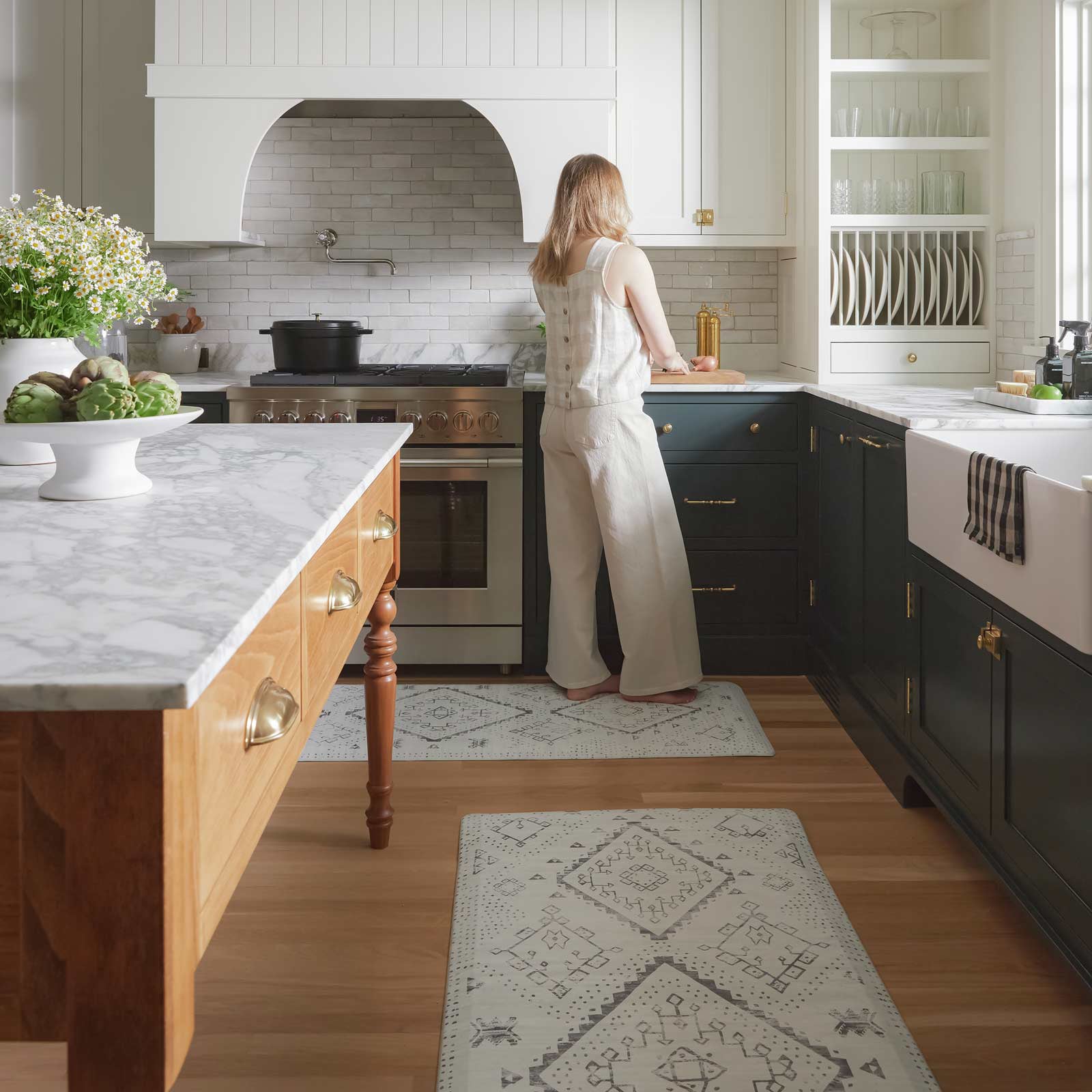 Ula avalon black and white boho print standing mat shown in a kitchen in sizes 30x72 and 22x72 in between the counter and island with a woman standing on at the back counter using a cutting board