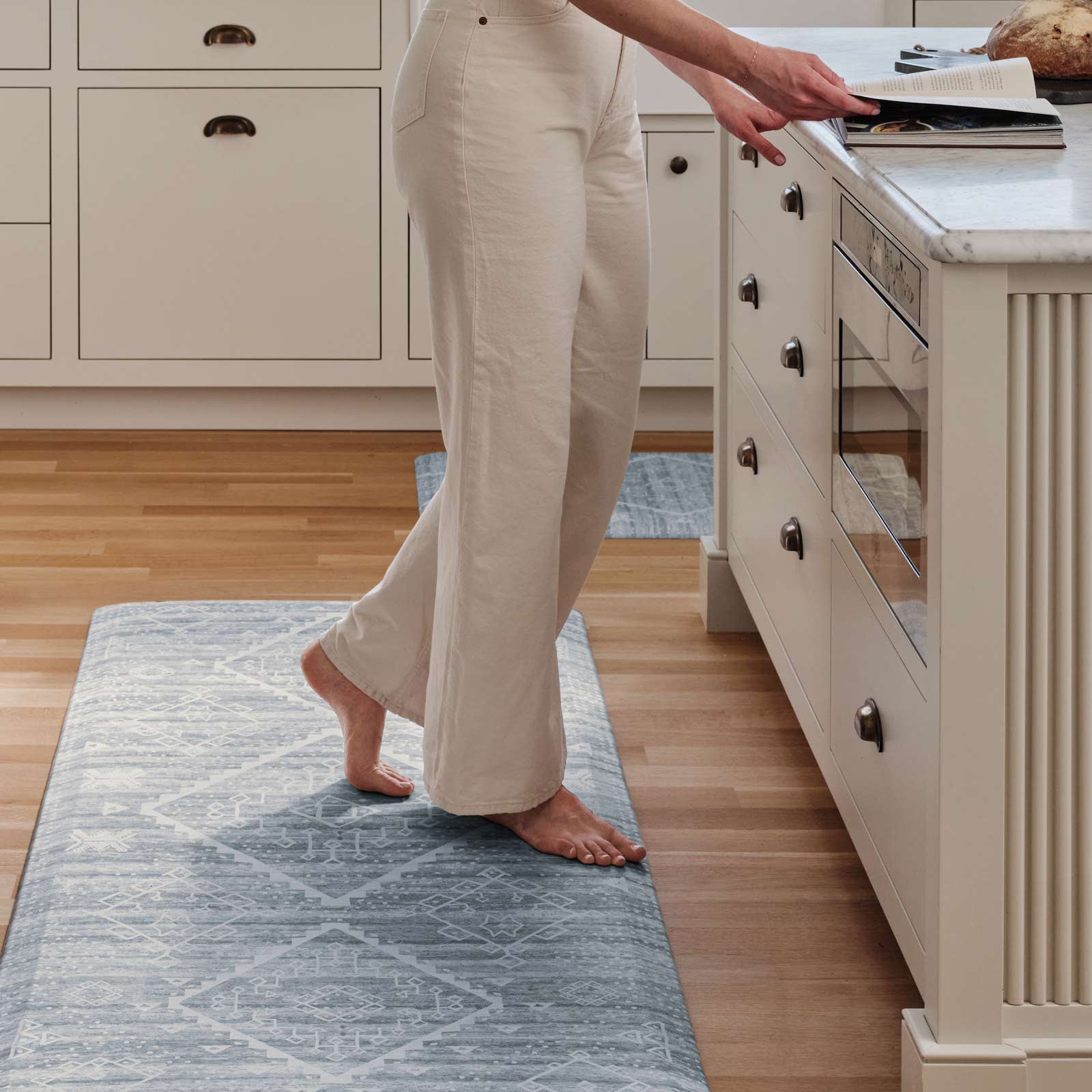 Ula blue and white boho print standing mat shown in kitchen in size 30x72 in front of the counter with woman standing on the mat reading a cook book