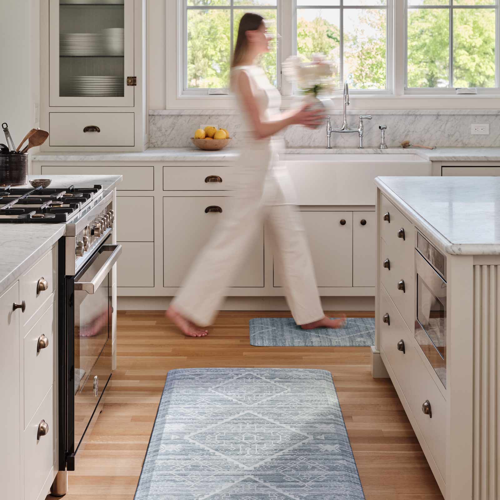 Ula blue and white boho print standing mat shown in kitchen with size 30x72 in between the island and counter and size 22x54 in front of the sink with woman walking by carrying flowers