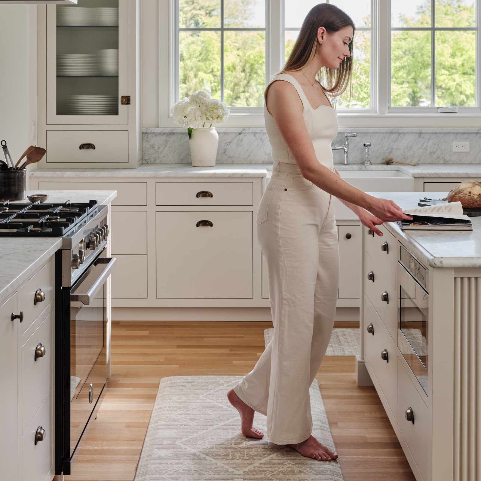 Ula driftwood neutral boho print standing mat in size 30x72, shown in a kitchen with a woman standing on the mat at the kitchen counter reading a cookbook