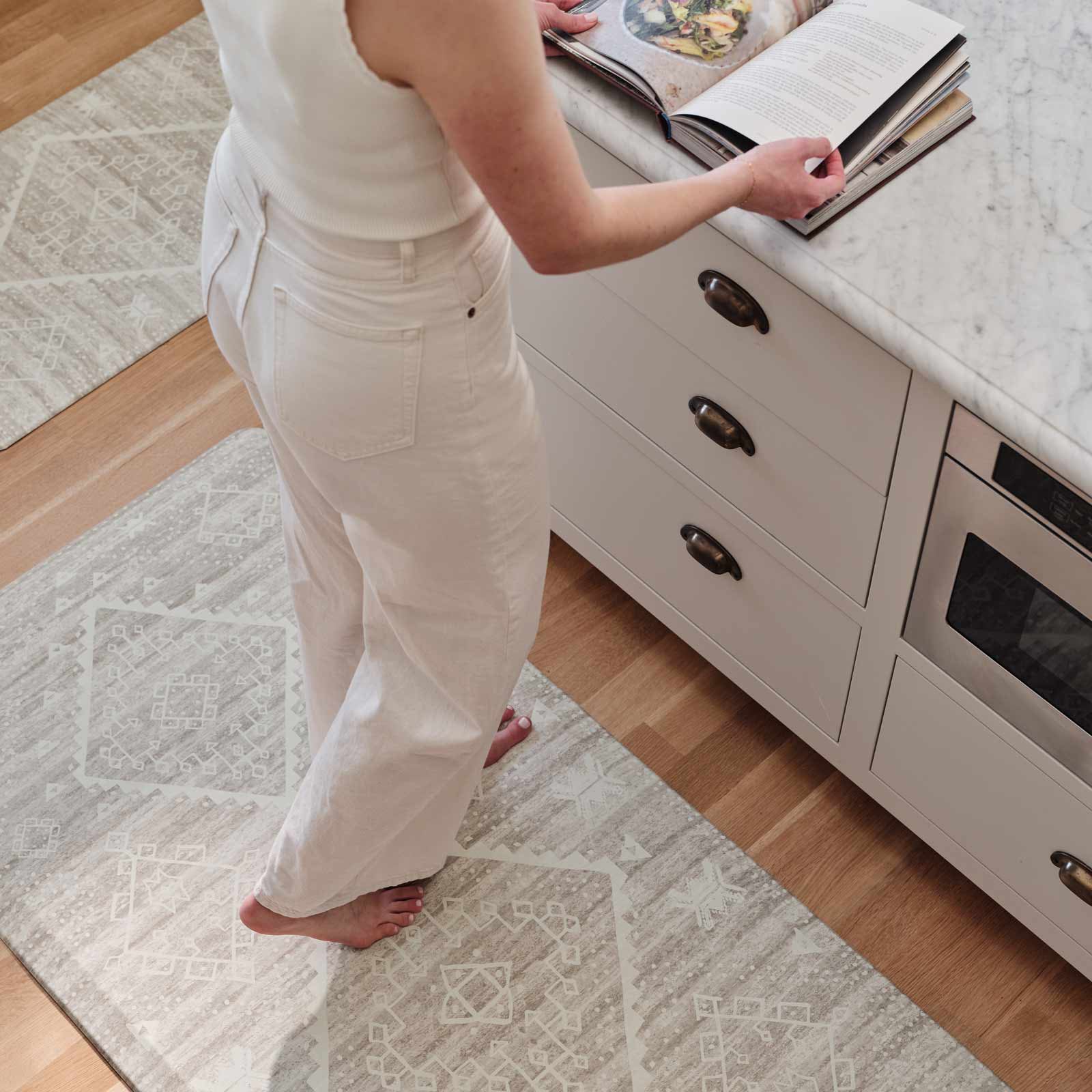 Ula driftwood neutral boho print standing mat shown in a kitchen with size 22x72 and 30x72 on either both sizes of a kitchen island with a woman standing on the 30x72 reading a cookbook