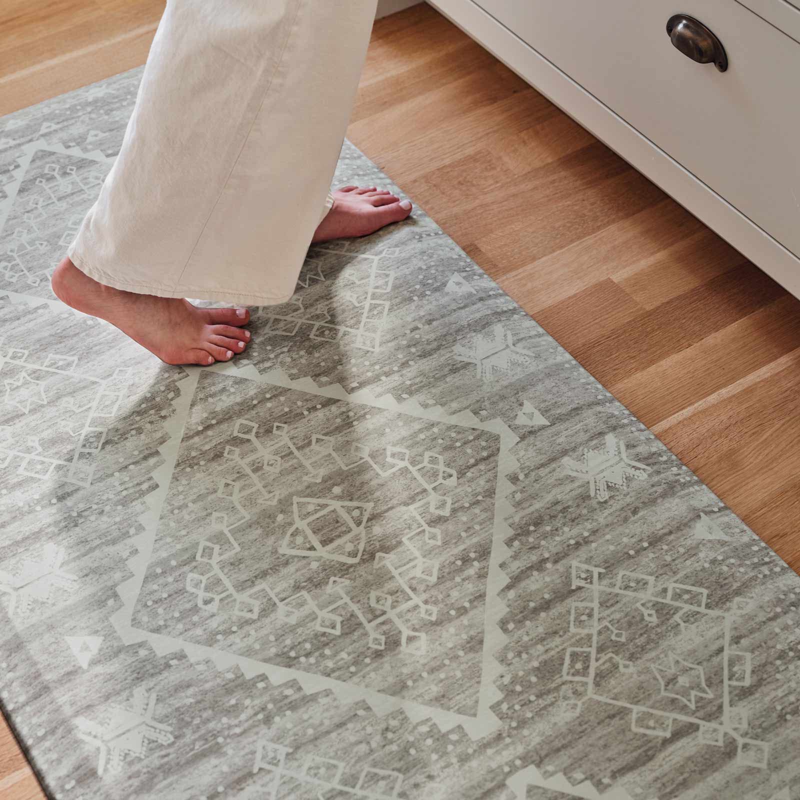 Ula Willow light green and white boho print standing mat on a wood floor in front of a kitchen counter shown close up with womans feet standing on the mat