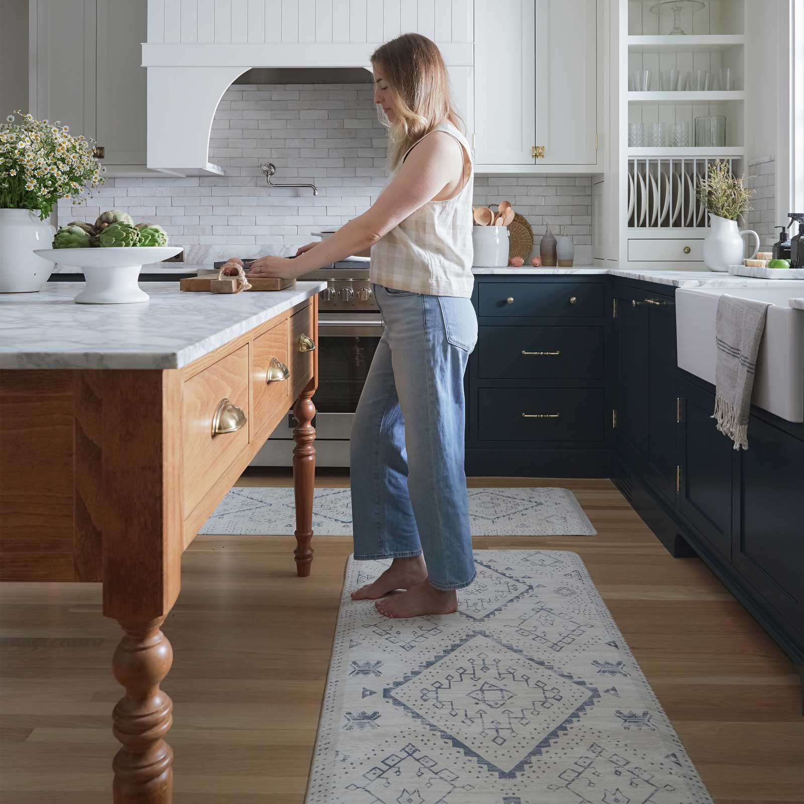 Ula Windmill blue and white boho print standing mat shown in a kitchen with size 30x72 and 22x72 in between the island and counter and a woman standing on size 30x72 using a cutting board on the island