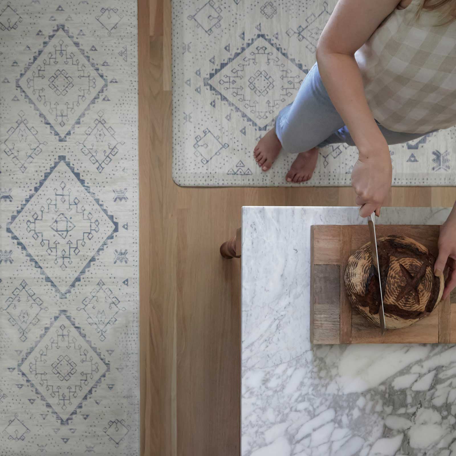 Overhead shot of the Ula Windmill blue and white boho print standing mat in 2 sizes around a kitchen counter with a woman standing on one of the mats cutting bread