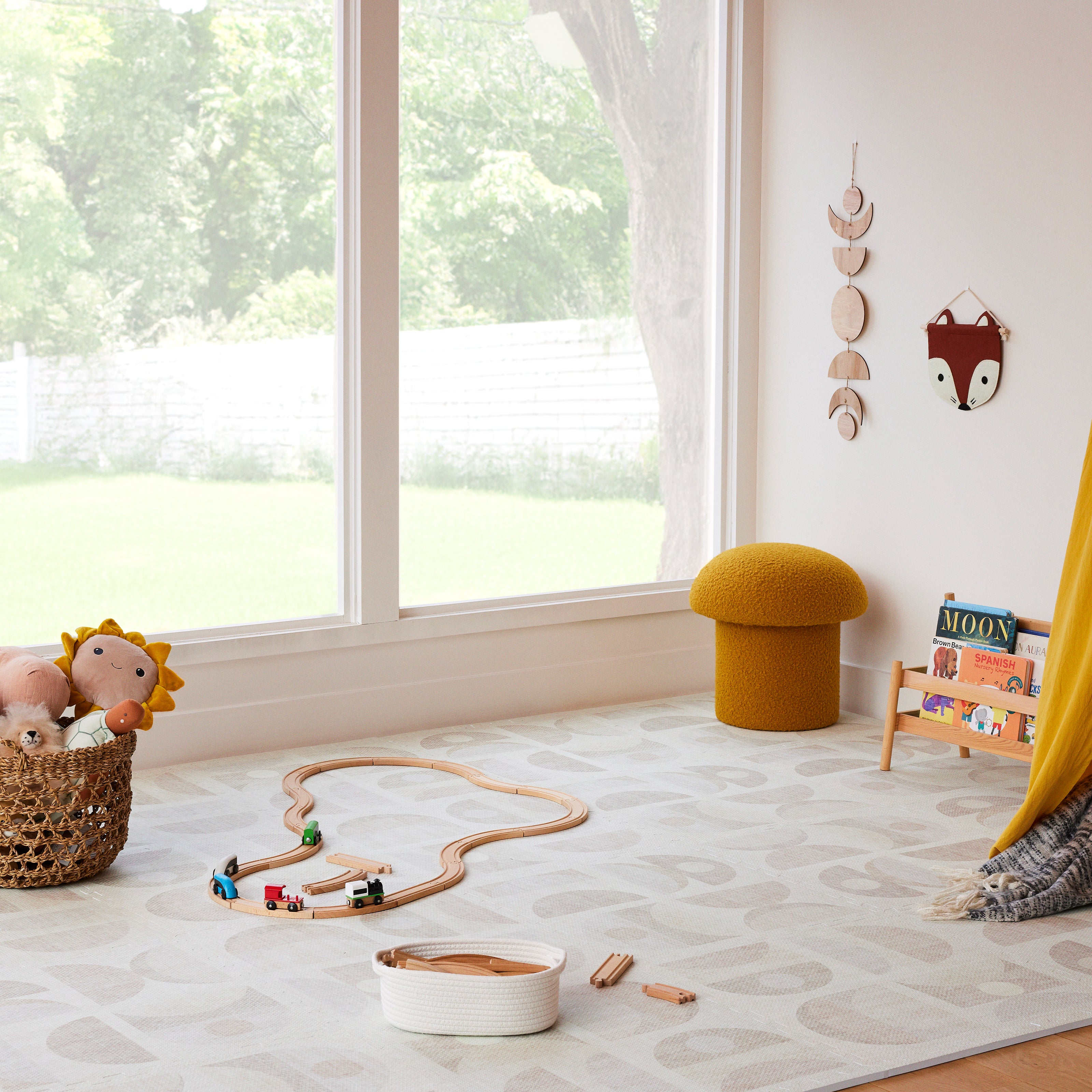 Luna sandstone neutral geometric print little nomad play mat in front of a big window in a play room with toy train tracks, stuffed animals, and book stand laid on the mat