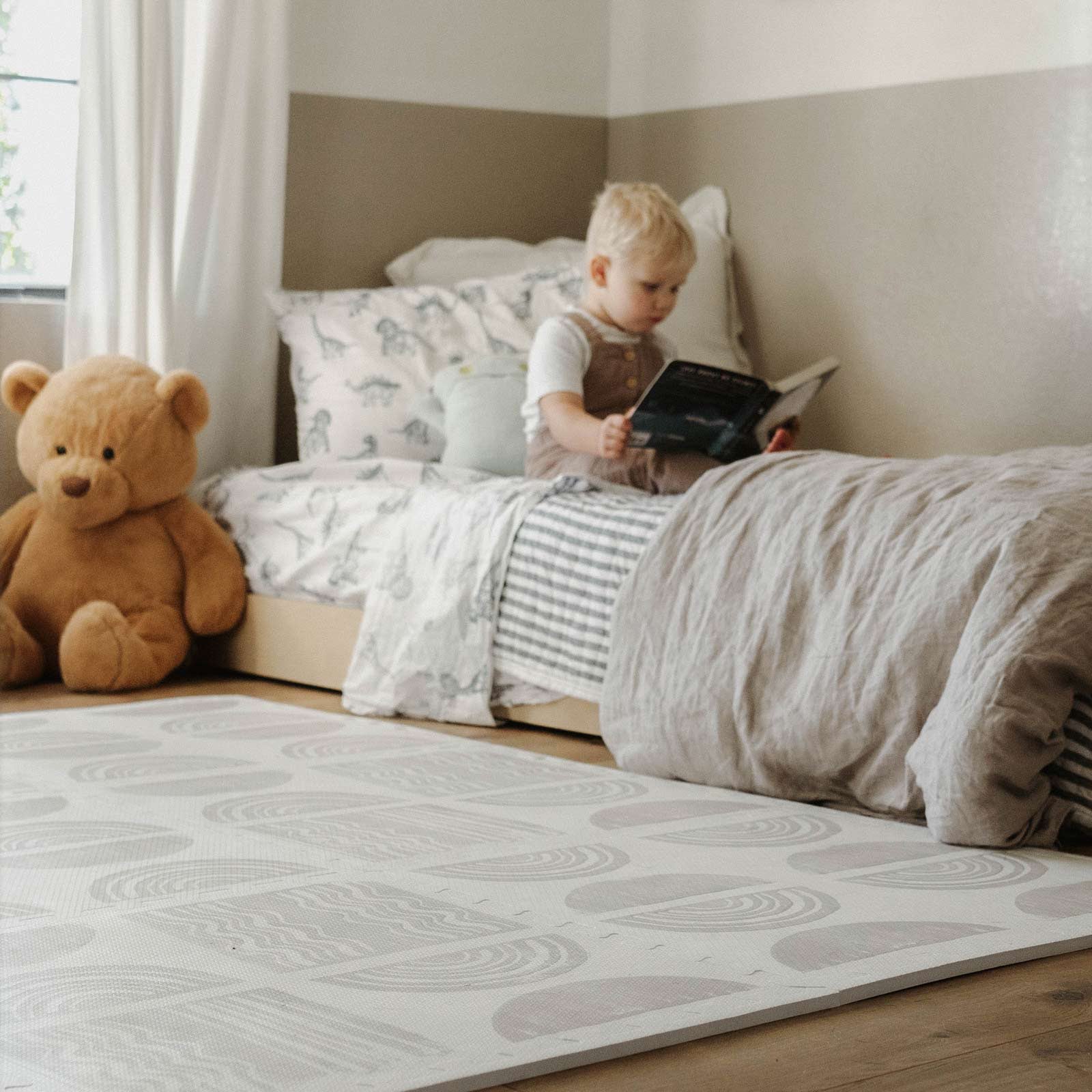 Ada pebble neutral block print play mat shown in bedroom next to a large stuffed teddy bear and toddler sitting on his bed reading a book. 