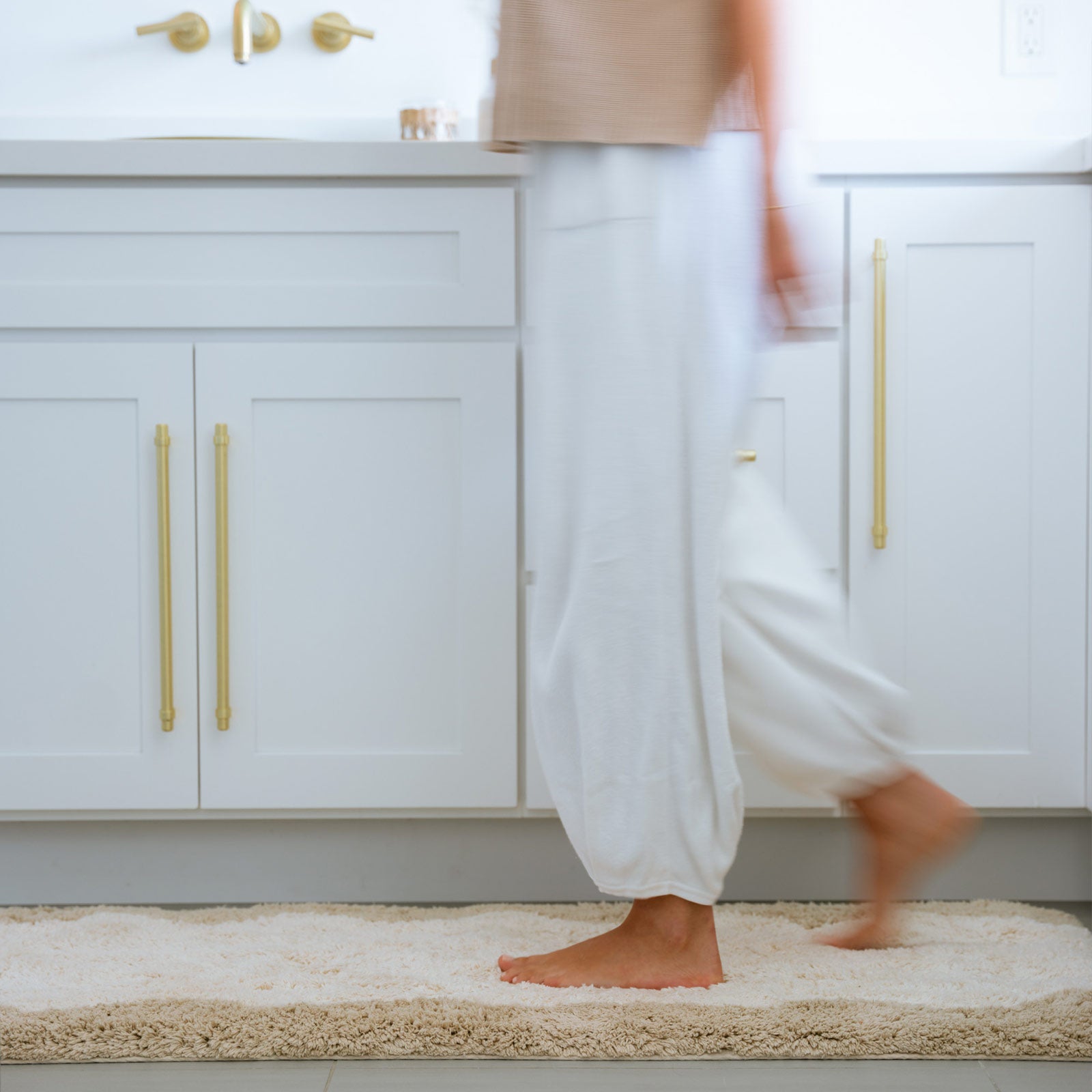 Harper au lait beige scallop print bath mat shown in front of a vanity with woman walking across size 24x70