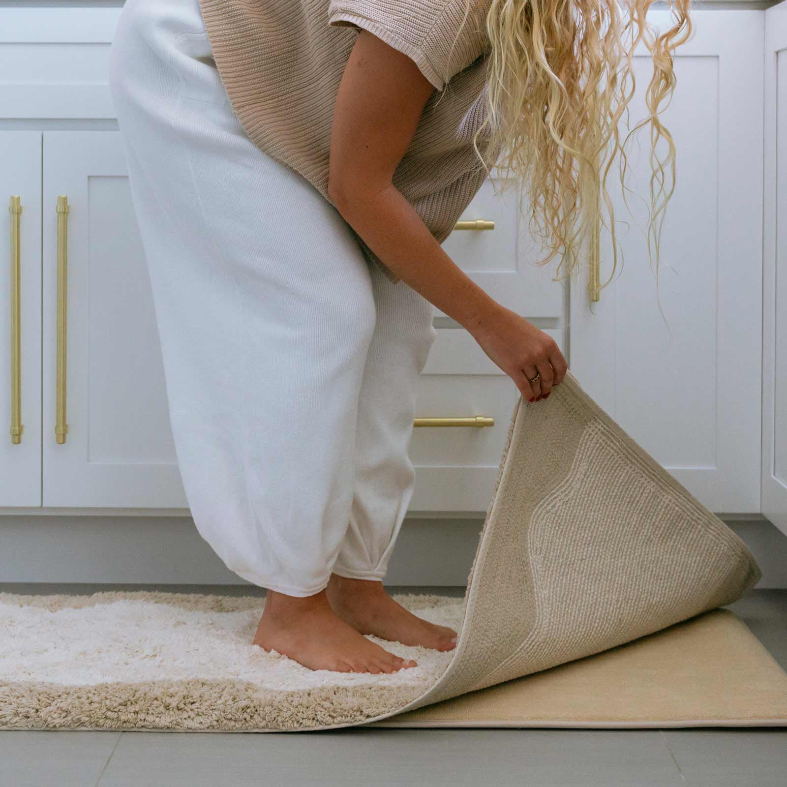 Harper au lait beige scallop print bath mat shown in front of a vanity in size 21x58 with woman standing on the mat lifting up the corner to show the memory foam liner underneath