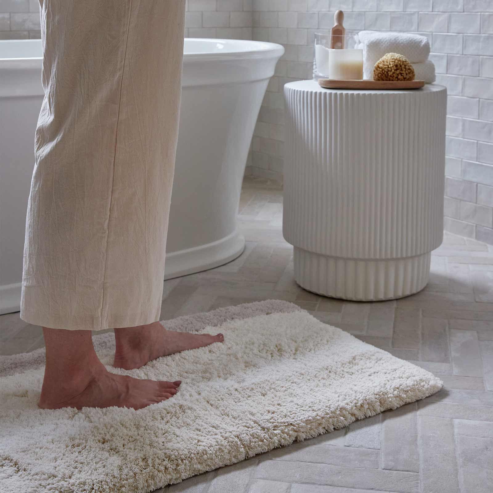 Riley Swan gray white and beige stripe print bath mat in size 21x34 in front of a bath tub with woman's feet standing on the mat