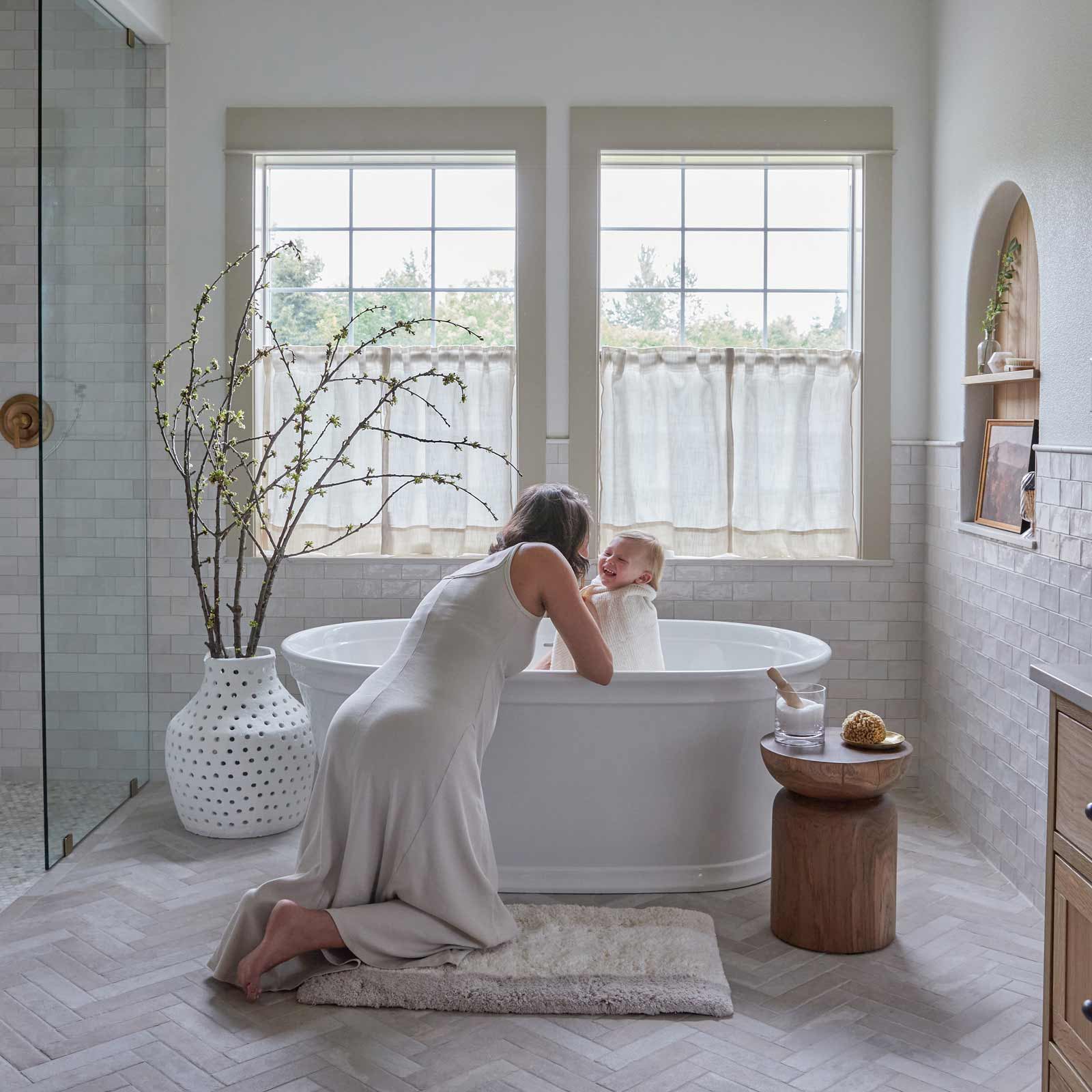 Riley Swan muted neutral stripe print bath rug in size 21x34 in front of a bath tub with woman kneeling on the mat while giving her baby a bath