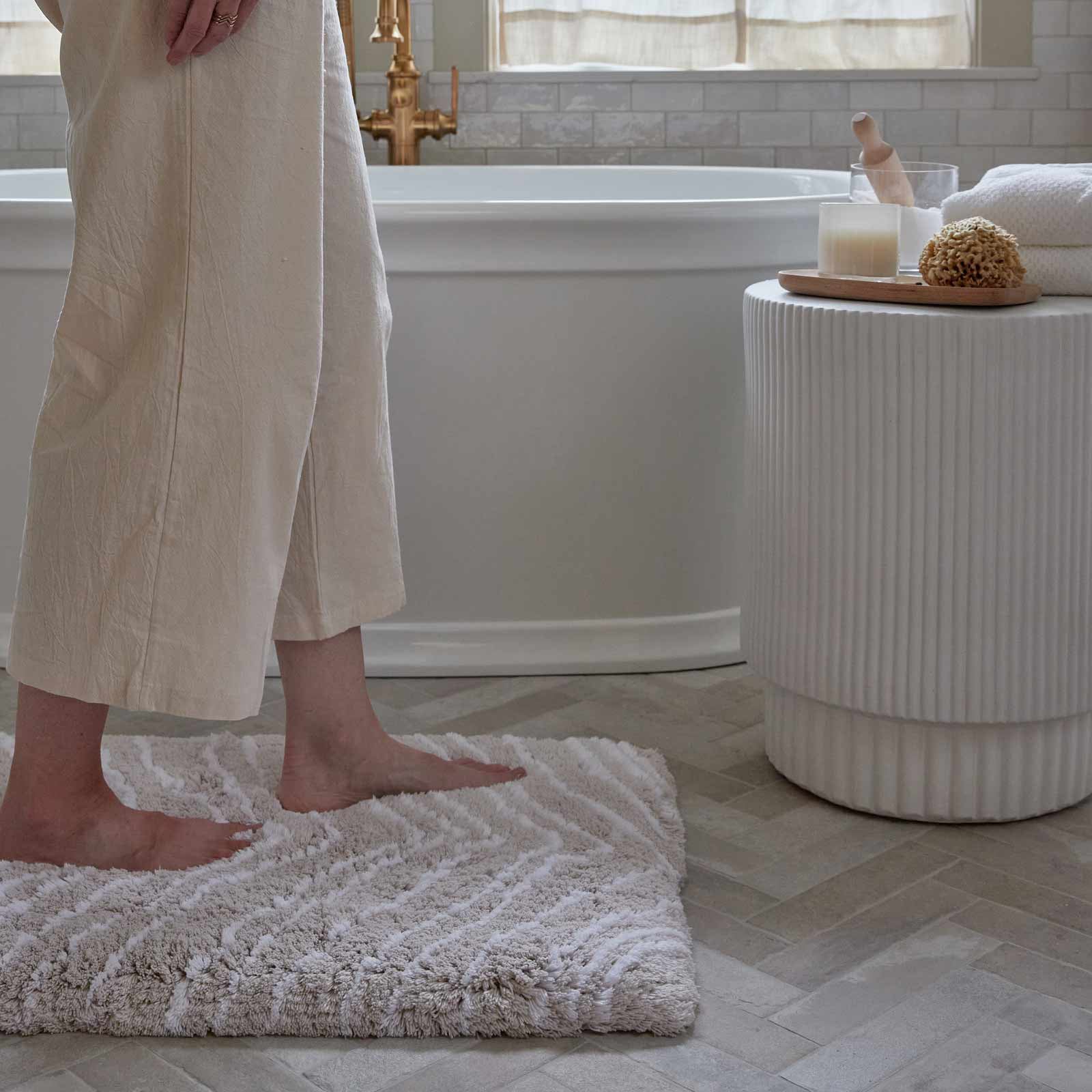 Zelda shell neutral zebra print bath rug shown in size 21x34 with woman's feet standing on the mat in front of a tub