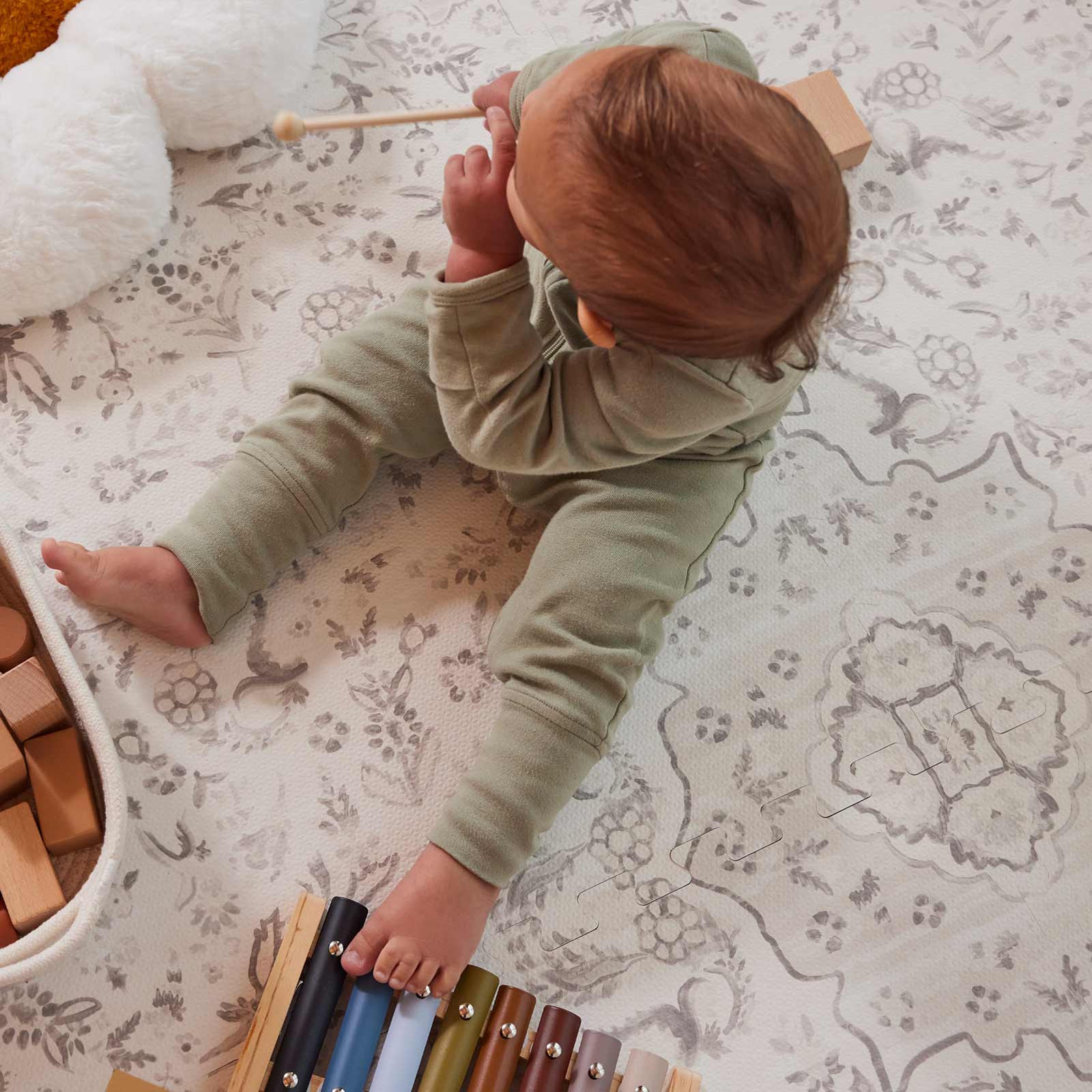 Overhead shot of the Emile Latte neutral floral little nomad play mat with baby boy sitting on the mat playing with a toy xylophone