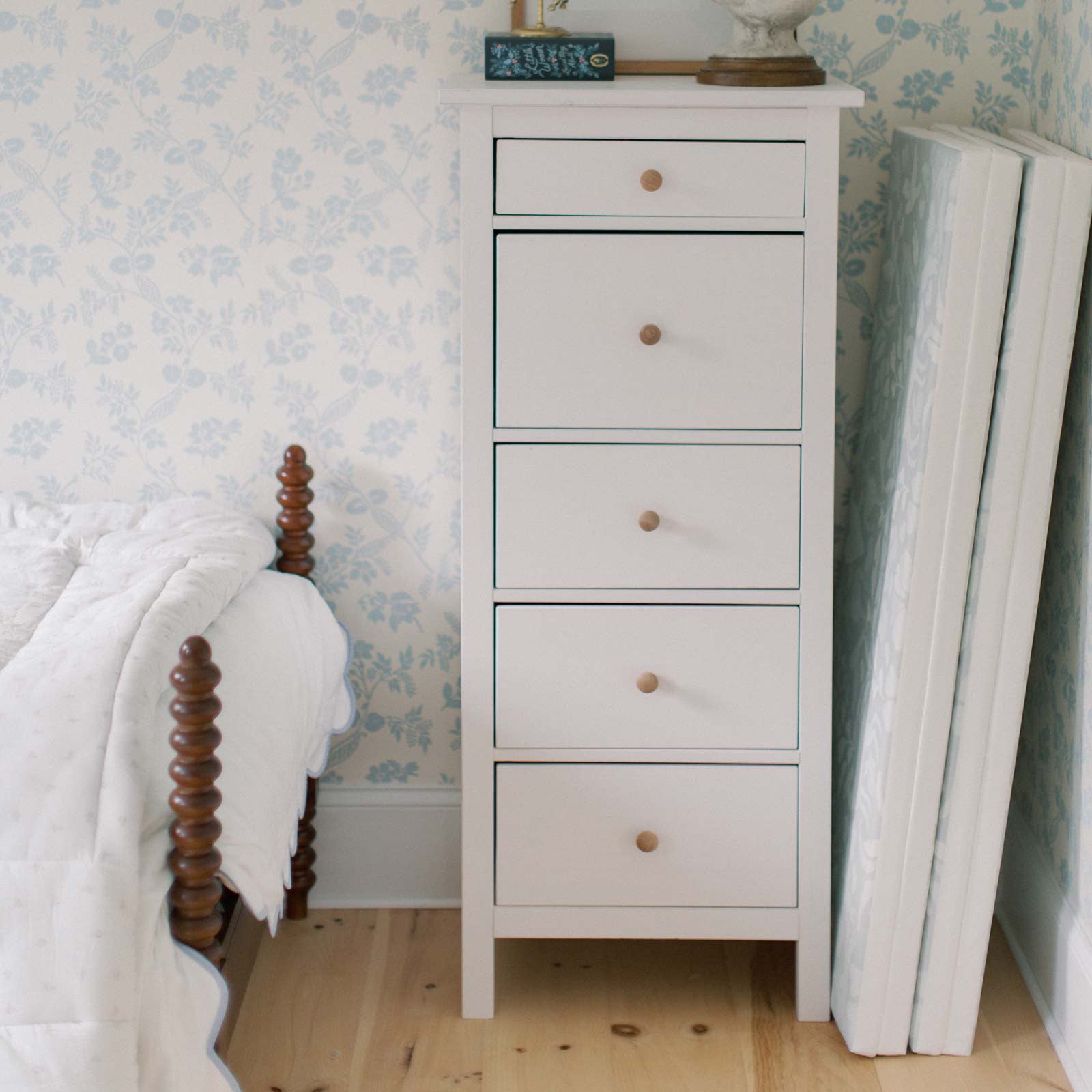 Bedroom with white blue floral wallpaper, the edge of a twin bed and tall white dresser with the Folklore Fog gray and white storybook pattern tumbling mat folded up and tucked into the space between the dresser and wall