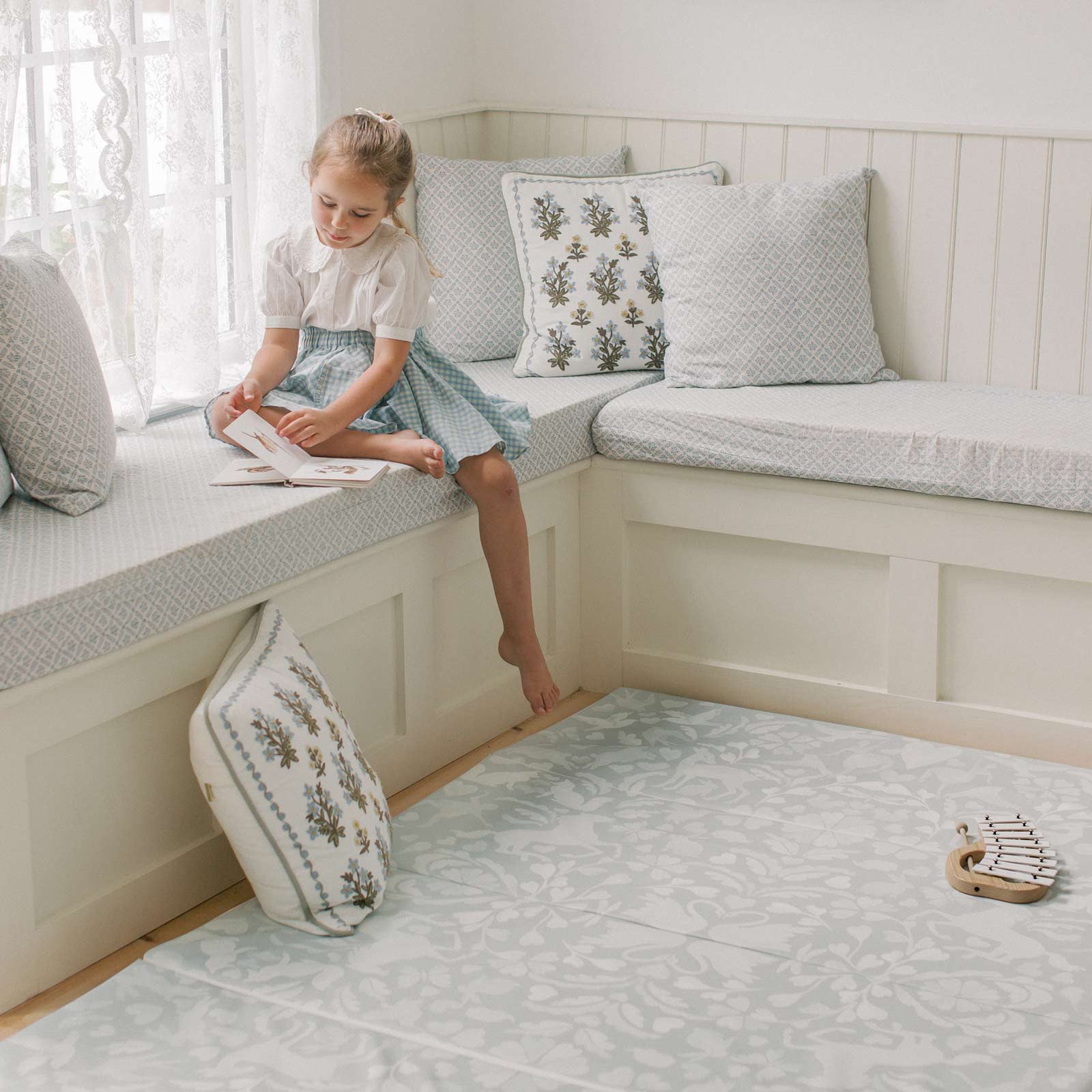 Folklore fog gray and white storybook pattern tumbling mat shown in a corner nook of a living room with little girl sitting in the window seat reading a book