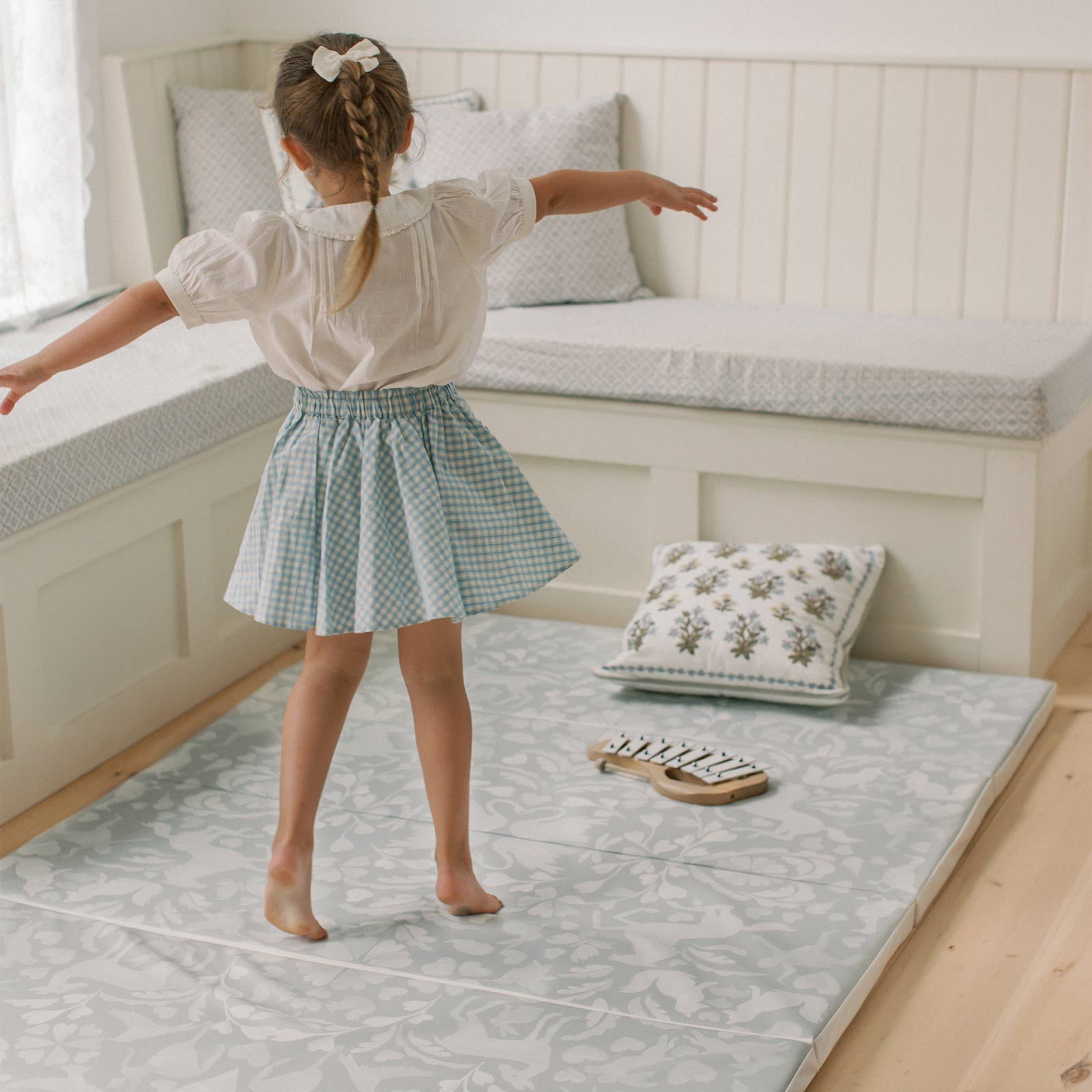 Folklore fog gray and white storybook pattern tumbling mat shown in a corner nook of a living room with little girl spinning around on the mat