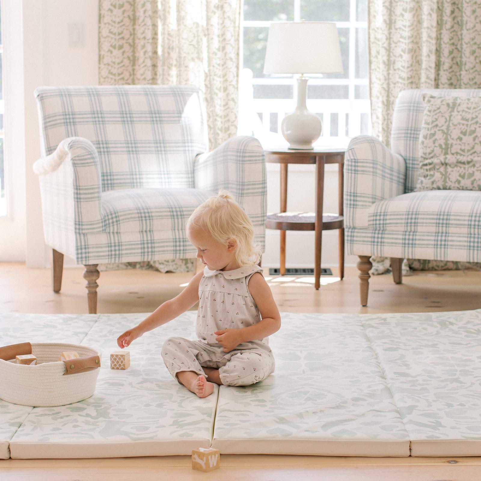 Folklore mint green storybook pattern tumbling mat shown in living room with toddler girl sitting on the mat reaching for a basket full of blocks