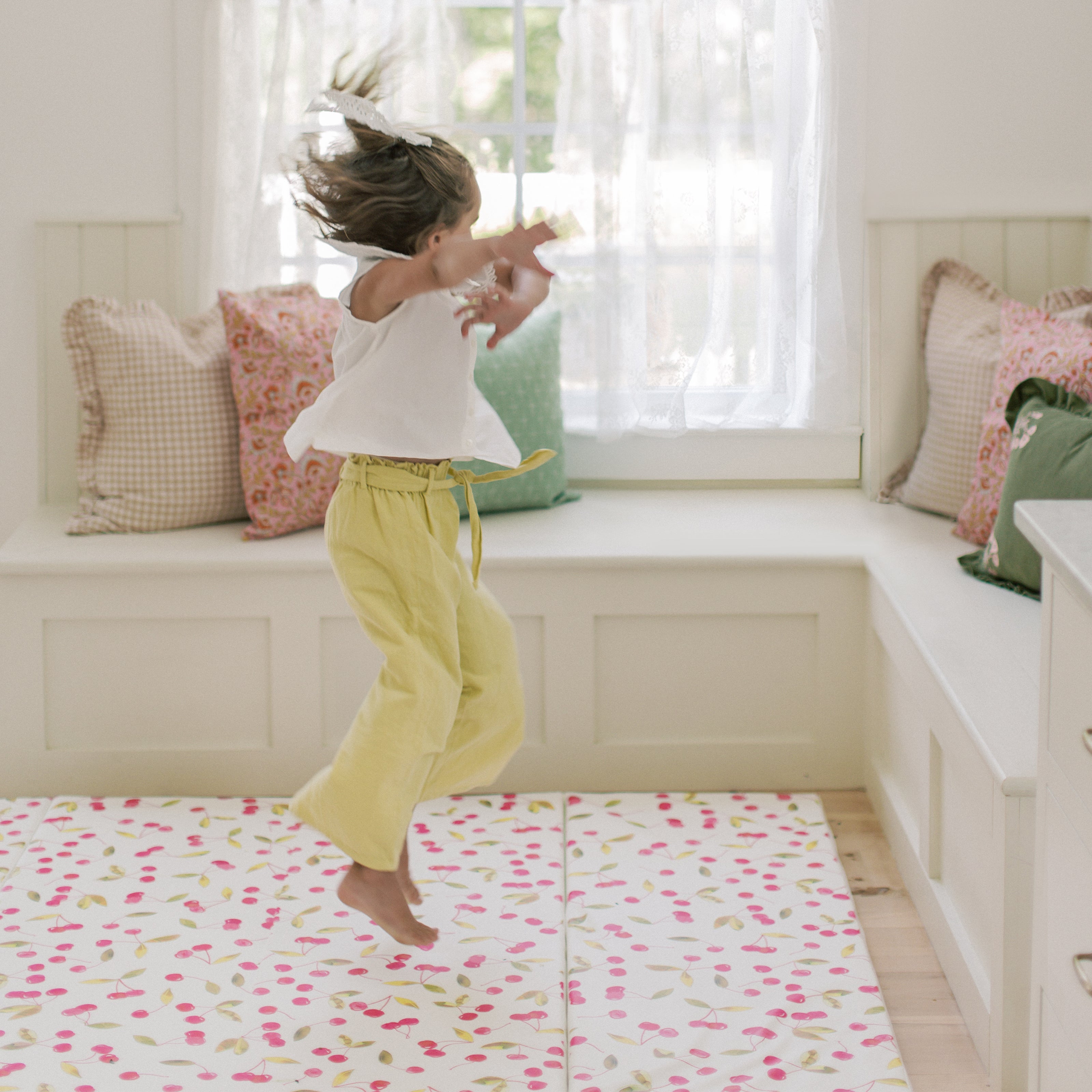 Lulu red and green Cherry print tumbling mat shown in living room with little girl jumping off of the mat into the air