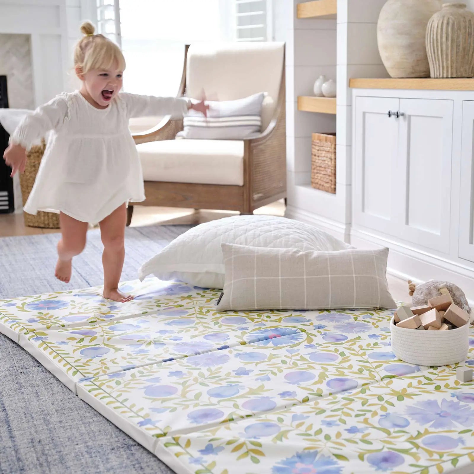 Bluebell blue green and white floral tumbling mat shown in living room with pillows and basket of wooden blocks sitting on the mat with toddler girl running across the mat