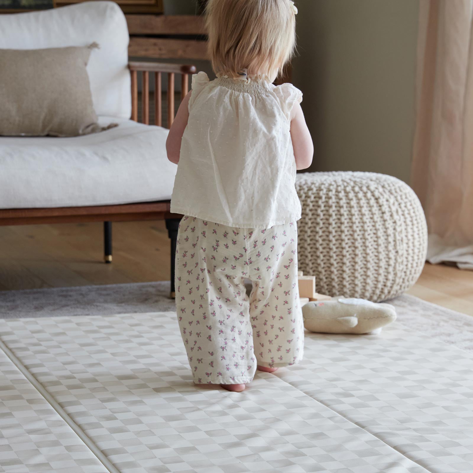 Almond neutral checker print tumbling mat shown in living room with baby girl walking across the mat toward toys and stuffed animals