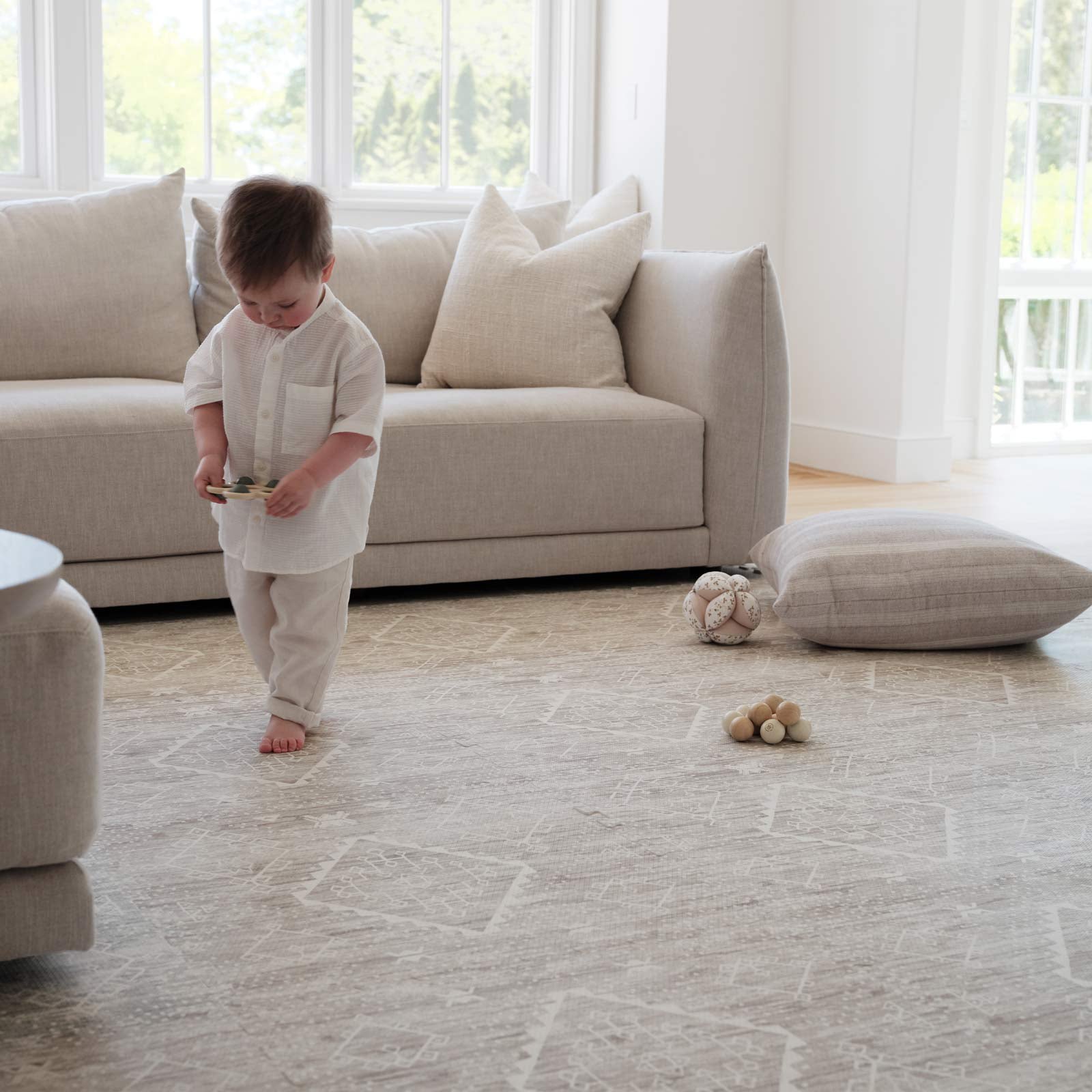 Ula driftwood neutral boho pattern play mat shown in a living room with light linen couch and little boy holding a toy with more toys and a pillow laid behind him on the mat