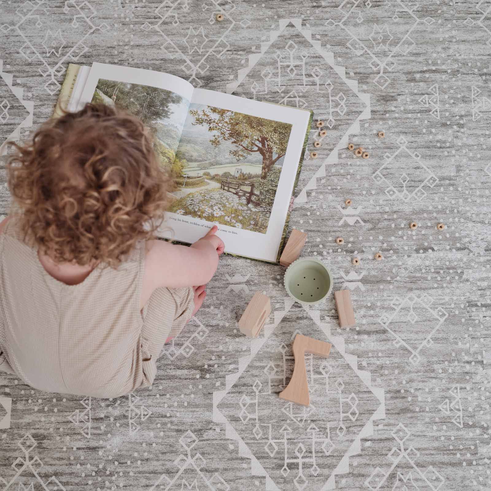 Ula willow washed green boho print play mat shown from above with toddler boy reading a book with wooden blocks and cheerios on the mat