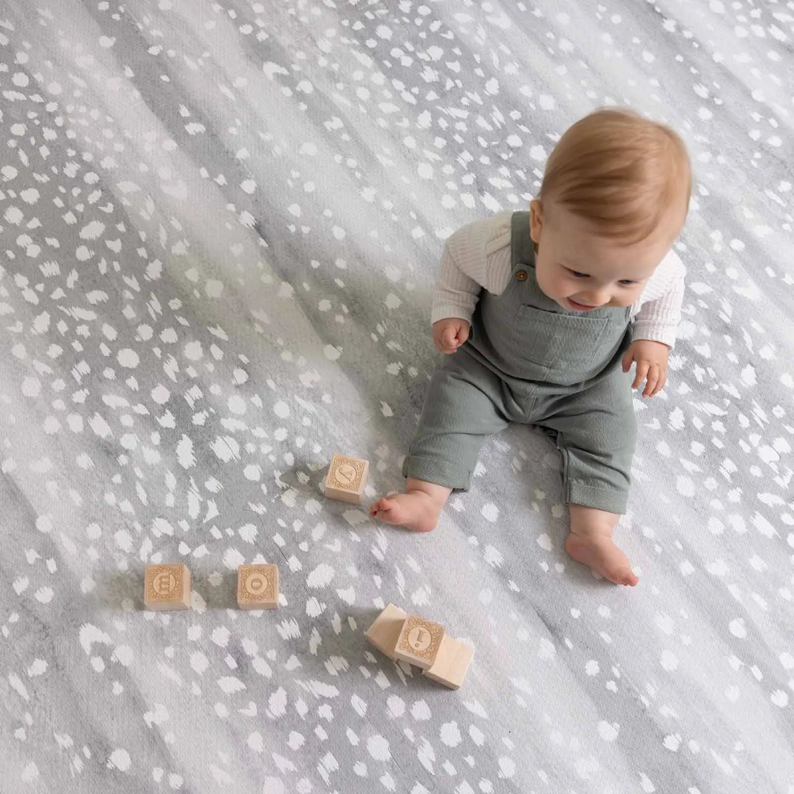 Little Nomad Play Mat in Fawn silver gray and white animal print. Shown from above with baby sitting on the mat next to wooden blocks