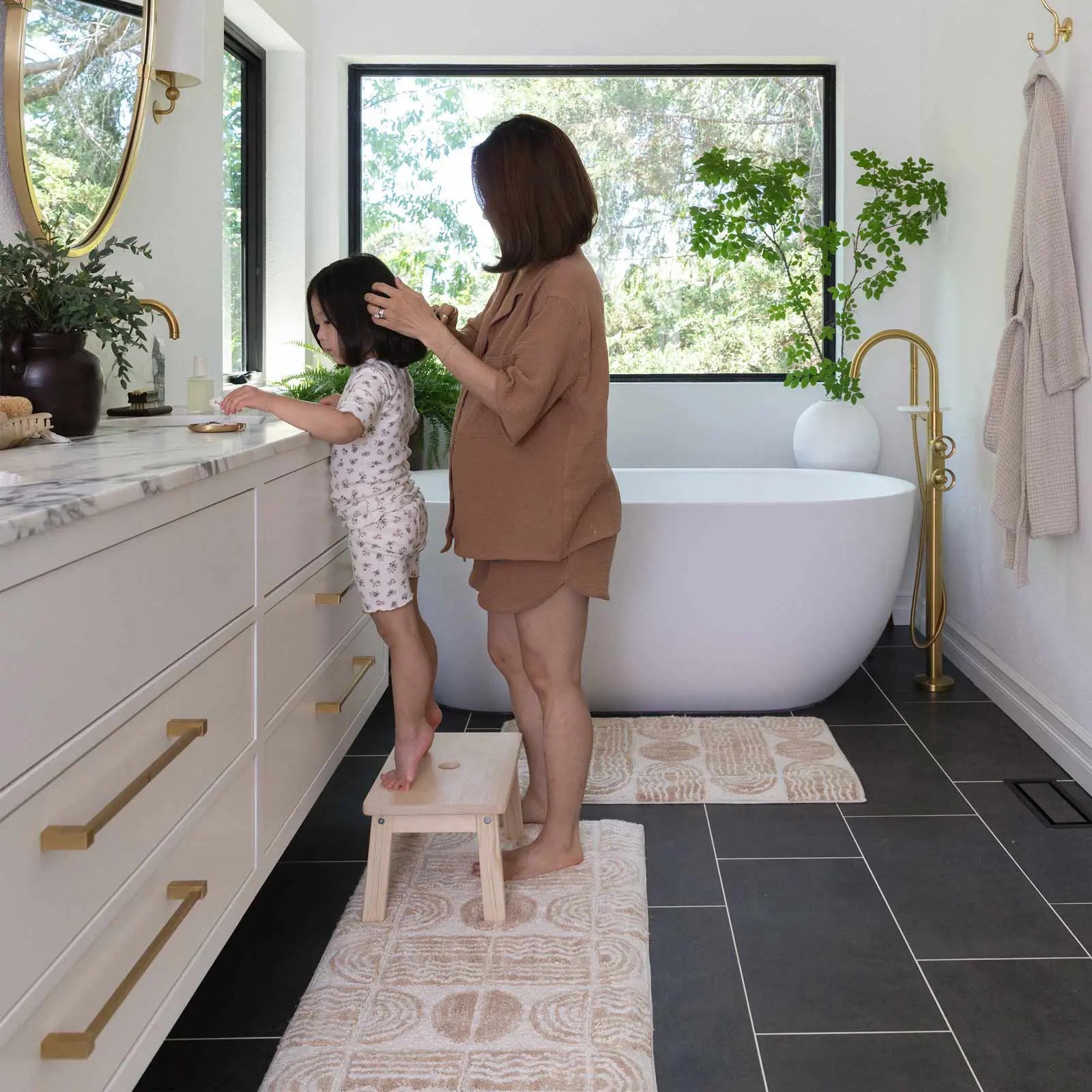 Bathroom with Ada Pebble neutral geometric print bath mat in size 21x58 in front of a double vanity and size 21x34 in front of a tub. Little girl standing on a stool in front of the vanity while her Mom is behind her doing her hair.