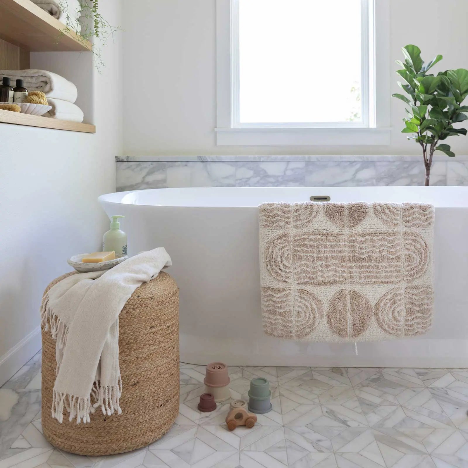 Ada Pebble neutral and white geometric print bath mat in size 21x34 in a bathroom hanging over the edge of a tub with bath accessories laid out on a stool next to the tub