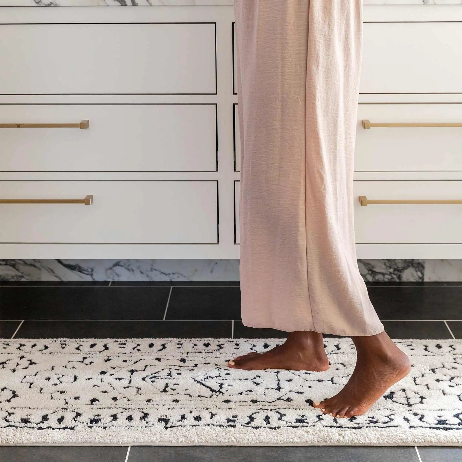 Arden noir black and white persian rug pattern bath mat in size 21x58 in front of a bathroom vanity with close up shot of a woman's legs standing on the mat