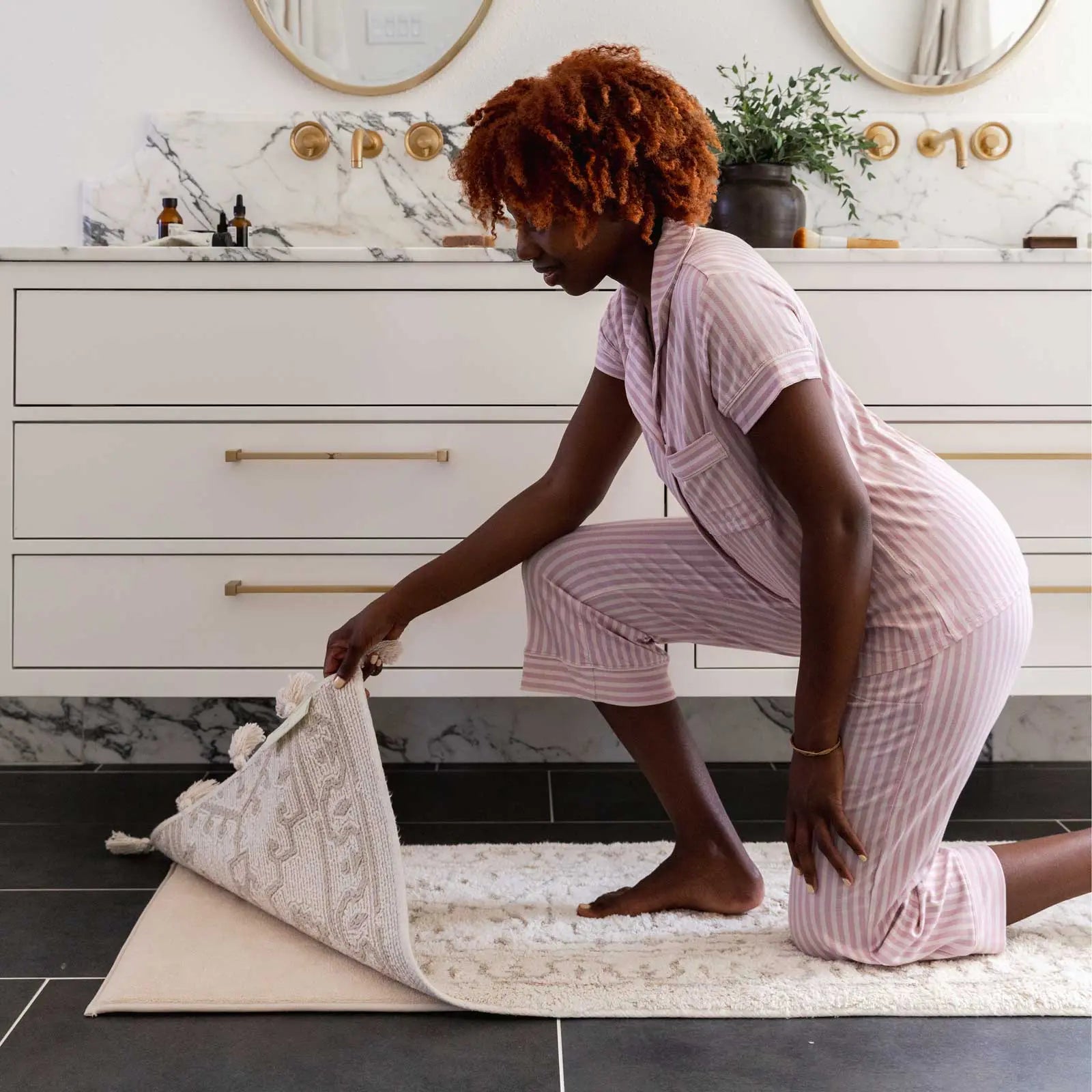 Arden pearl cream and white persian rug pattern bath mat in size 21x58 in front of a bathroom vanity with woman kneeling on the mat and lifting the corner to show the foam liner