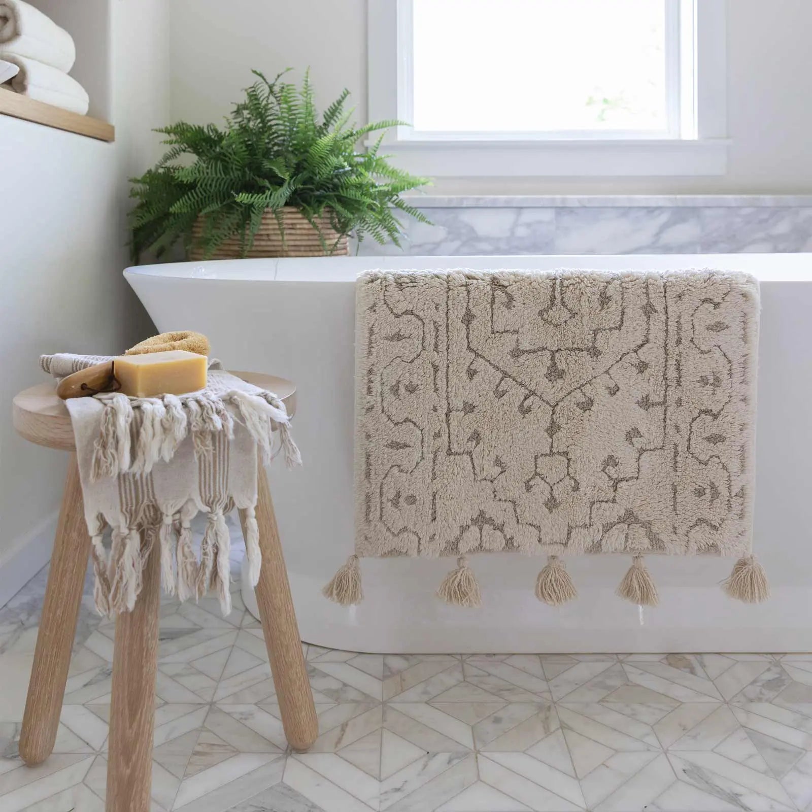 Arden pearl cream and white persian rug pattern bath mat in size 21x34 shown hung over the side of a bathtub and next to the tub theres a wooden stool holding bath goods