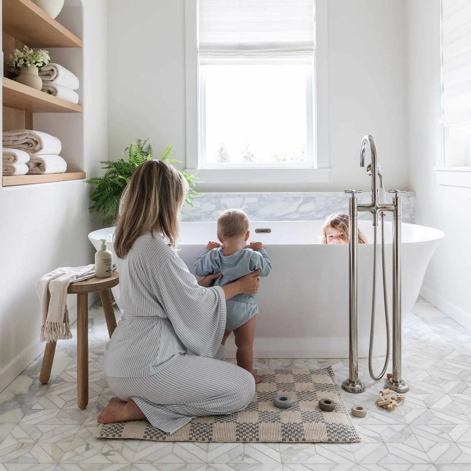 Bath mat in checker harbor gray print in size 21x34 shown in a bathroom in front of a tub with Mom sitting on the mat supporting baby holding itself up on the tub