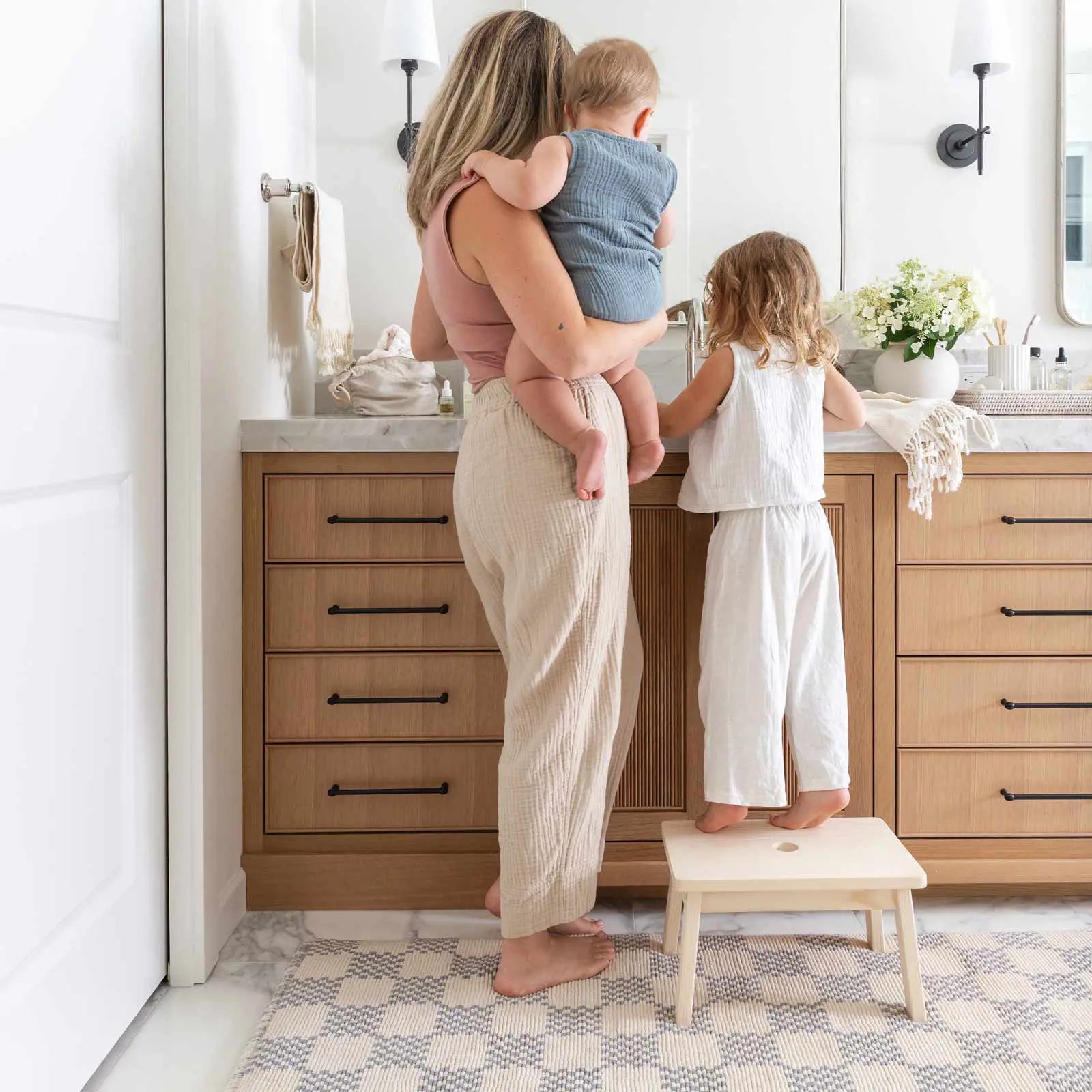 Bath mat in checker harbor gray print in size 21x58 in front of a double vanity with Mom holding a baby and toddler on a stool on the mat