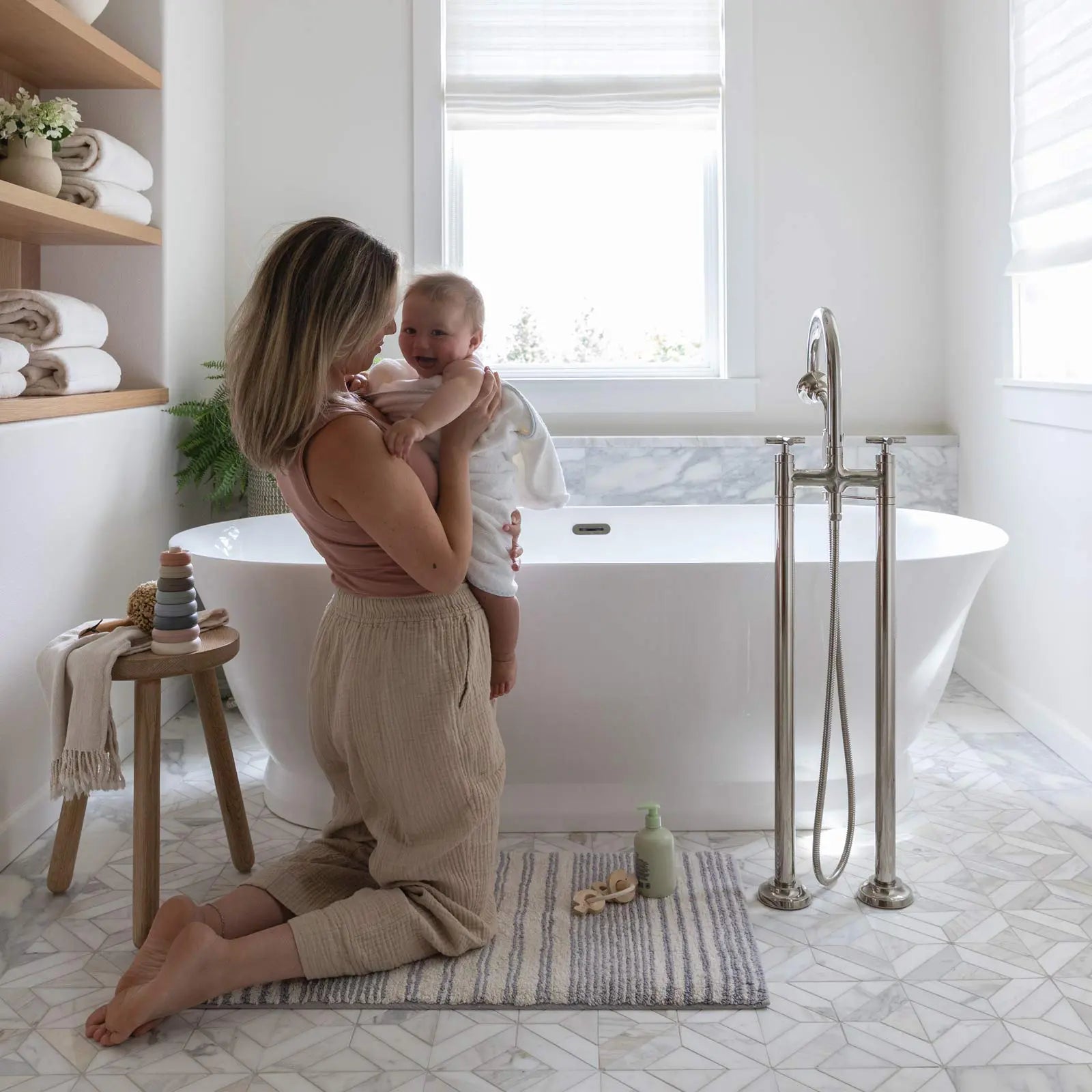 Coastal Nantucket blue and white stripe bath mat in size 21x34 shown in a bathroom in front of a tub with Mom kneeling on the mat hugging baby after a bath