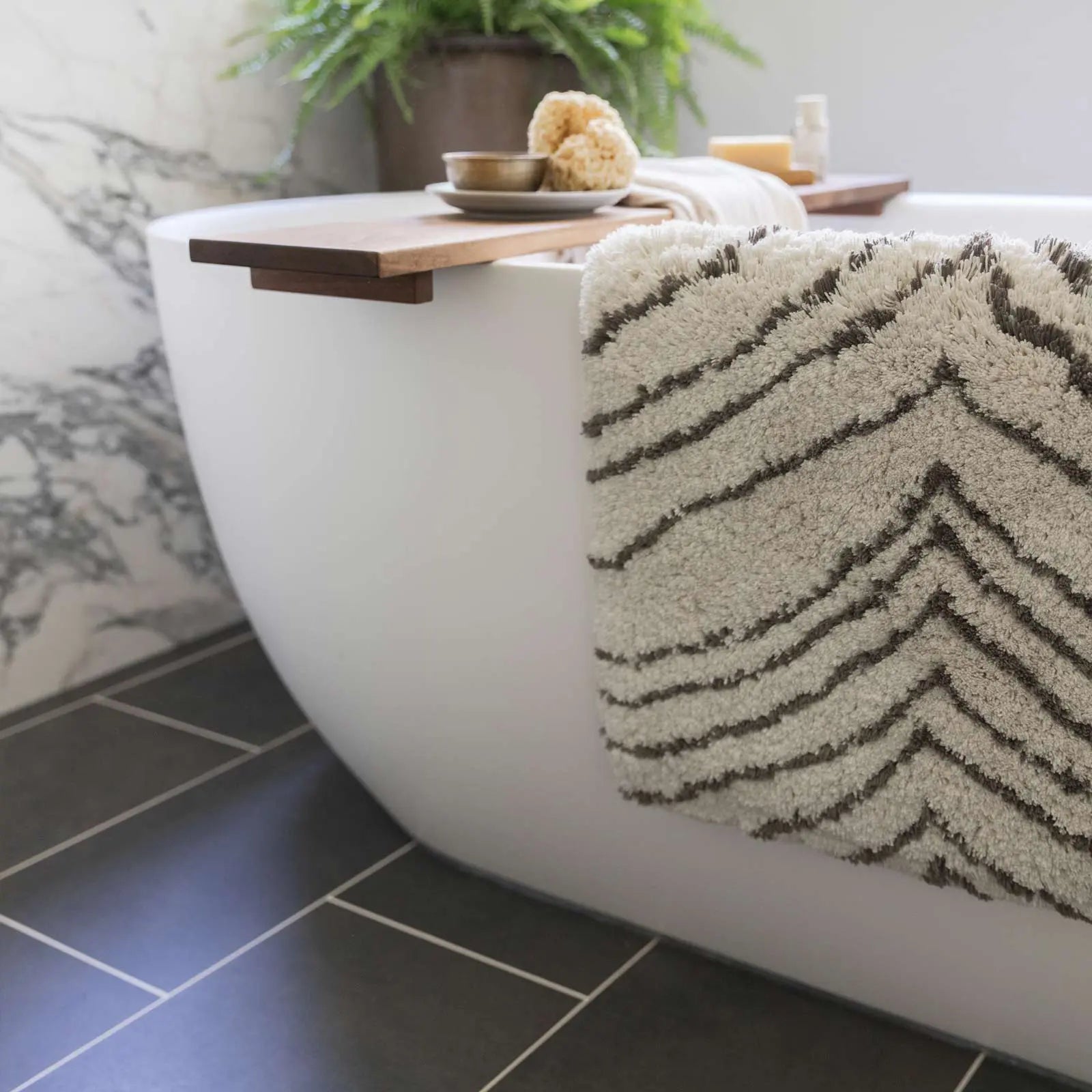 Zelda puffin cream and gray zebra print bath mat in size 21x34 shown hanging over the edge of a bathtub with a wooden spa table across the tub