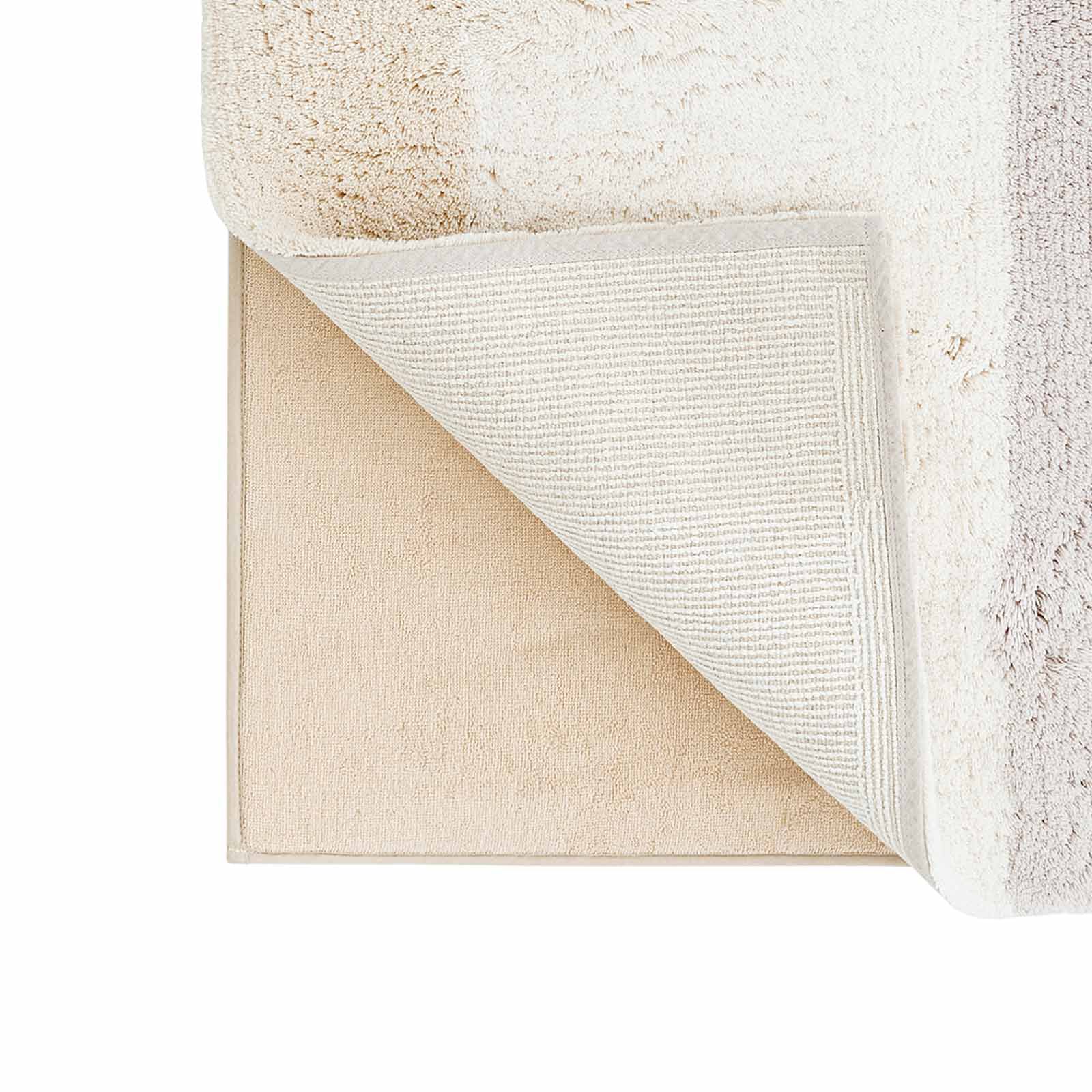 Corner of the Riley Swan gray white and beige stripe print bath mat lifted up to show the foam liner underneath