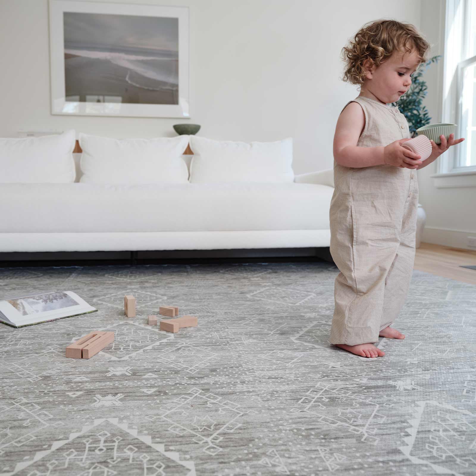 Ula willow washed green boho play mat shown in a living room with toddler boy standing on the mat holding toys with white sofa, book, and wooden blocks behind him